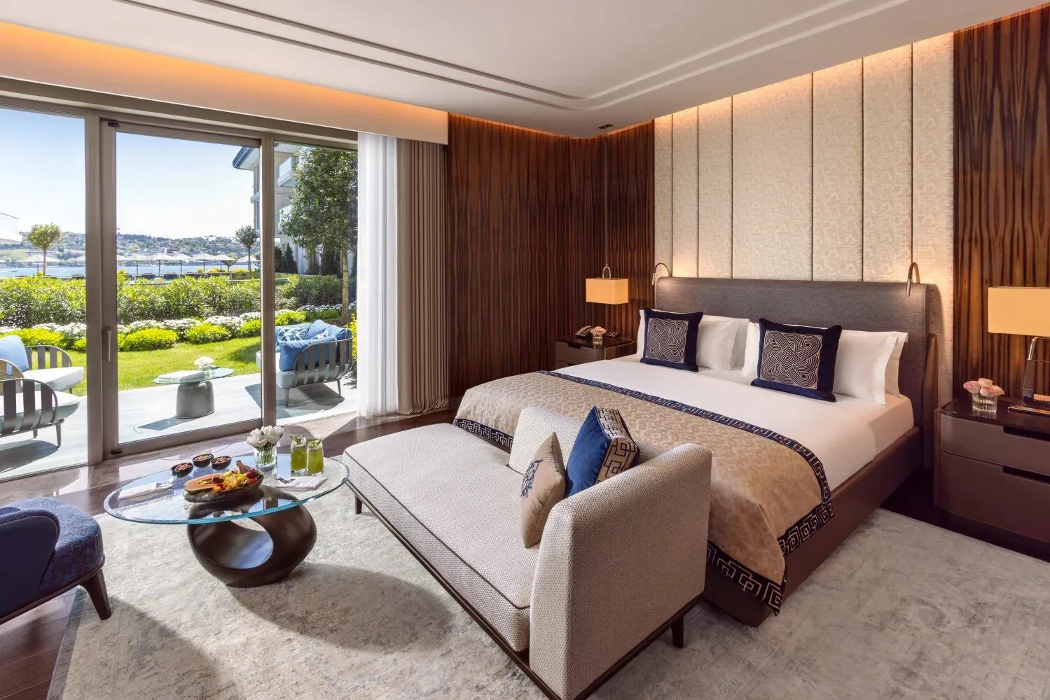 Suite de hotel con cama king, paneles de madera de ébano, chaise lounge en bouclé y vista al jardín con terraza